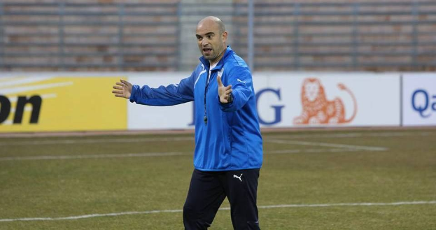 Ein Mann in einer blauen Jacke steht entschlossen auf einem Fußballfeld, umgeben von Tafeln mit Text und Logos während eines Trainings.