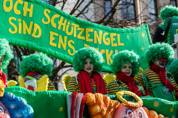 Eine Gruppe von Clowns in bunten Kostümen marschiert, hält ein Banner mit Text, mit Bäumen und einem Gebäude im Hintergrund.