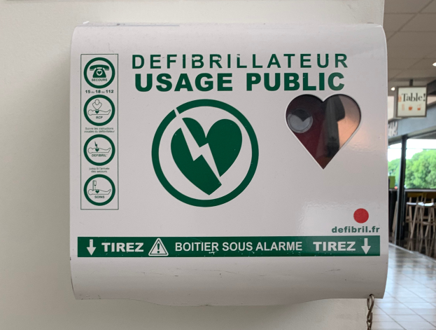 Ein Defibrillator-Gebrauchs-Schild an einer Gebäudewand, umgeben von Stühlen, Tischen, Brettern, Lampen und Bäumen im Hintergrund und dem Boden unten.