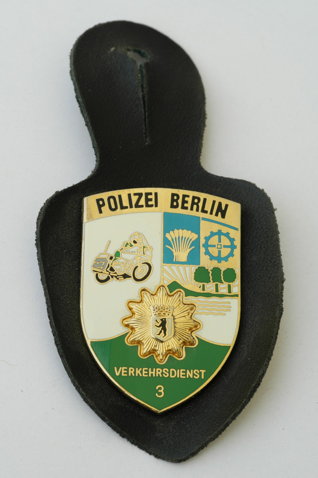 Ein schildförmiges Polizeisiegel mit 'Polizei Berlin' in fettem schwarzen Text auf einem blauen Hintergrund mit einem weißen Stern und Rahmen, das auf einer Oberfläche ruht.