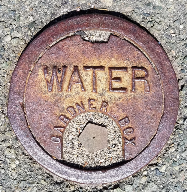 Ein verrosteter Metall-Schachtobjekt auf dem Boden mit dem Wort "wasser" darauf.