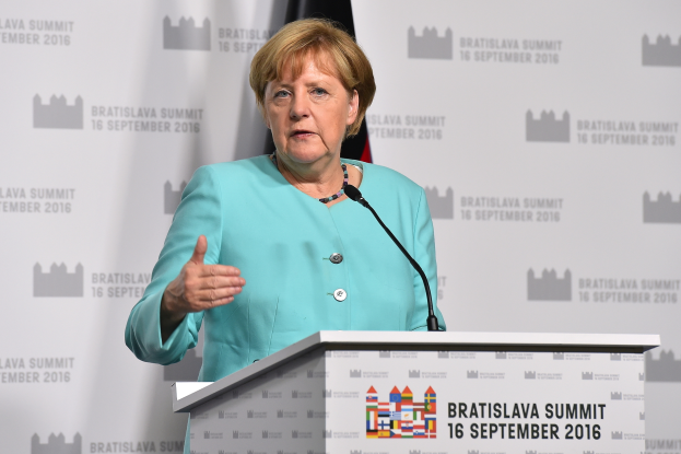 Bundeskanzlerin Angela Merkel hält eine Rede auf dem Bratislava-Gipfel, trägt ein blaues Kleid und steht vor einem Podium mit zwei Mikrofonen, mit einem Banner und einer Flagge dahinter.