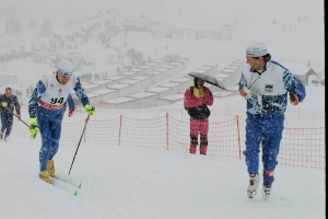 Eine Gruppe von Menschen, die eine schneebedeckte Piste hinunterskifahren, jeder hält einen Schirm, mit Hütten und Bäumen im Hintergrund.