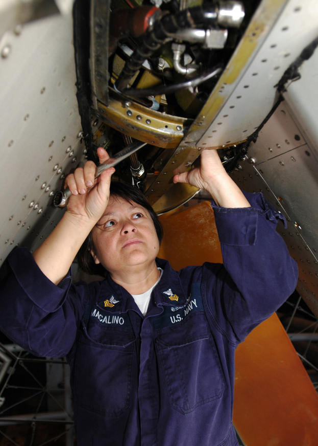 Eine Frau in blauer Uniform, die ein Werkzeug hält, arbeitet an einem Flugzeug und sieht konzentriert auf ihre Aufgabe.