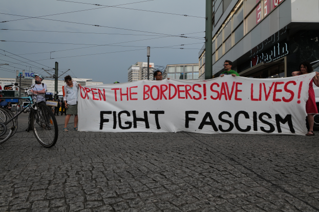 Eine Gruppe von Menschen auf der Straße mit einem Banner, auf dem 'Öffnet die Grenzen, rettet Leben, kämpft gegen Faschismus' steht, mit Fahrrädern auf der linken Seite, Gebäuden, Polen, Drähten und einem klaren blauen Himmel im Hintergrund.
