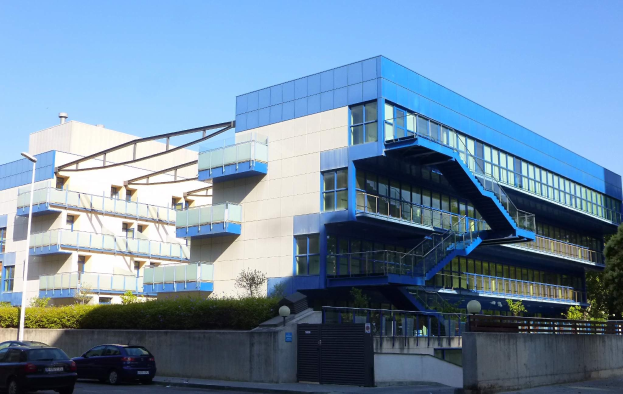 Großes modernes Gebäude mit zahlreichen parkenden Autos davor, umgeben von Straßenmasten, Lampen, Gittern, Grünflächen und einem klaren blauen Himmel, identifiziert als der neue Hauptsitz der University of California, Berkeley.