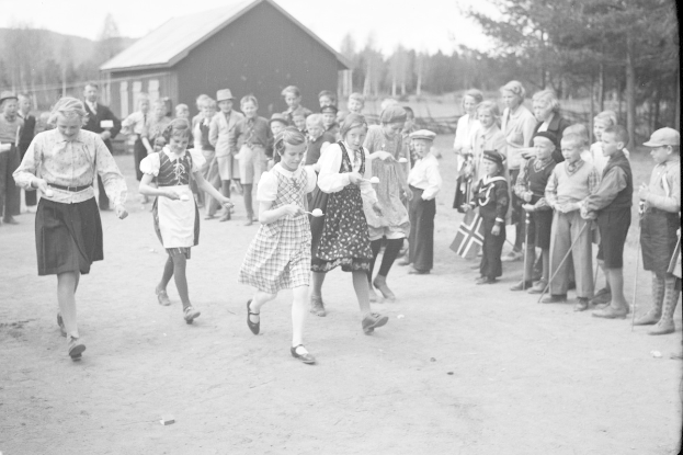 Schwarzes und weißes Foto von Kindern, die Fußball spielen, mit einer Menge Zuschauern, die Fahnen halten, Bäumen und einem Haus im Hintergrund.