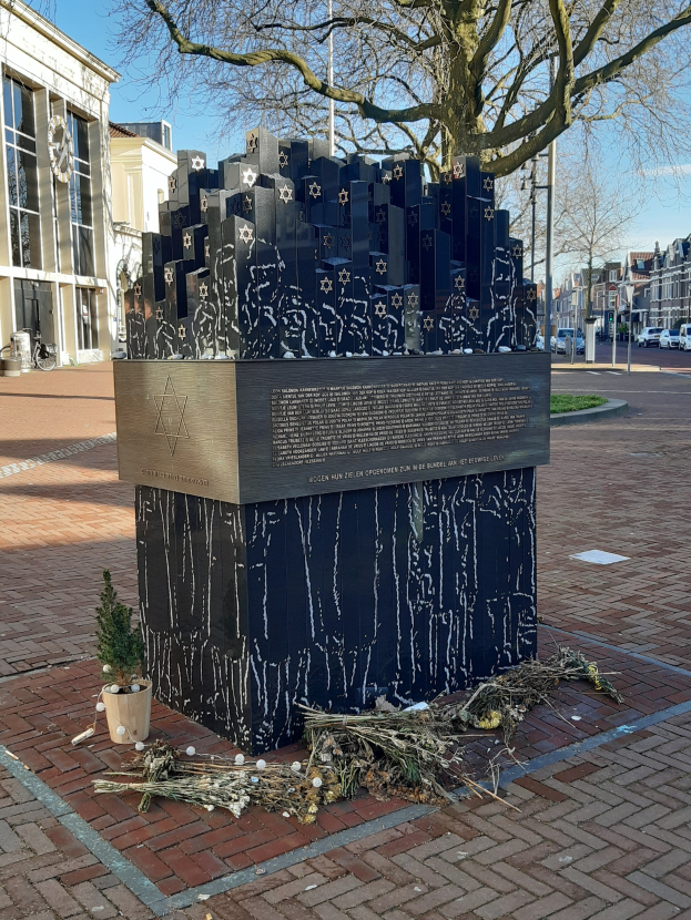 Denkmal in einem Stadtplatz, das an Holocaust-Opfer erinnert, mit einem Baum davor, einem Topfgewächshaus auf dem Boden, verstreuten trockenen Blättern und im Hintergrund Gebäude, Fahrzeuge und Bäume.