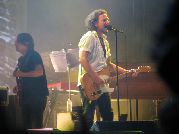 Ein Mann in einem weißen Hemd spielt Gitarre und singt in ein Mikrofon auf der Bühne, neben ihm ein anderer Mann in einem schwarzen Hemd, der ebenfalls Gitarre spielt; ein Piano, Kabel, Lautsprecher und Scheinwerfer sind im Hintergrund zu sehen.