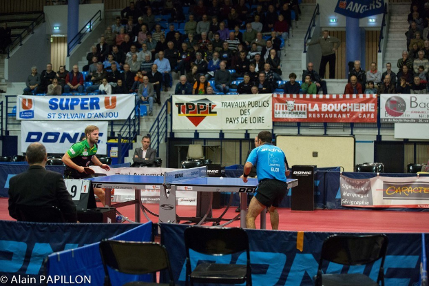 Indoor-Tischtennis-Match mit zwei Spielern im Wettbewerb, mehreren stehenden Zuschauern, sitzendem Publikum, leeren Stühlen und Werbetafeln in einer Stadionatmosphäre.