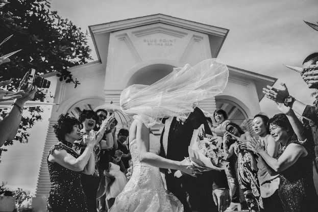 Ein Brautpaar steht in der Mitte, mit einem Gebäude im Hintergrund, einer Person mit einer Kamera links, einem Baum und einem Strauß, unter einem Himmel.