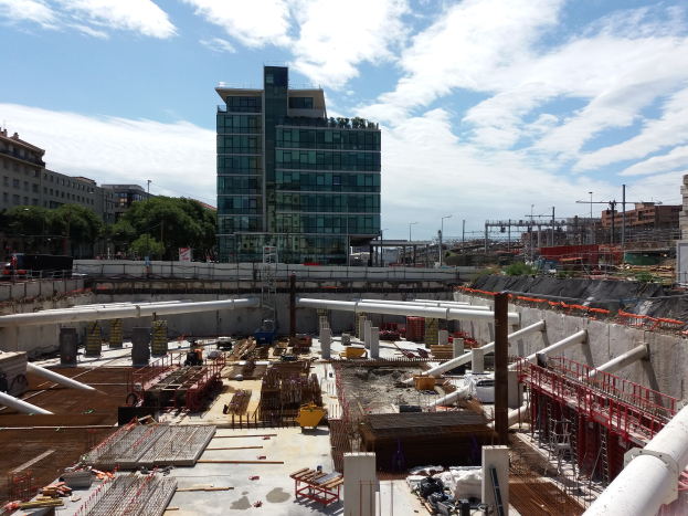 Eine Baustelle mit einem großen Gebäude, wahrscheinlich den Sitz der Europäischen Union, umgeben von Bäumen, Pfählen und Lichtern unter einem bewölkten Himmel.