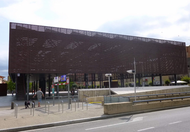 Großes modernes Gebäude mit zahlreichen Fenstern, das als neuer Eingang zur Stadt dient, umgeben von Straßeninfrastruktur, Fußgängern, Fahrzeugen und Bäumen unter einem bewölkten Himmel.