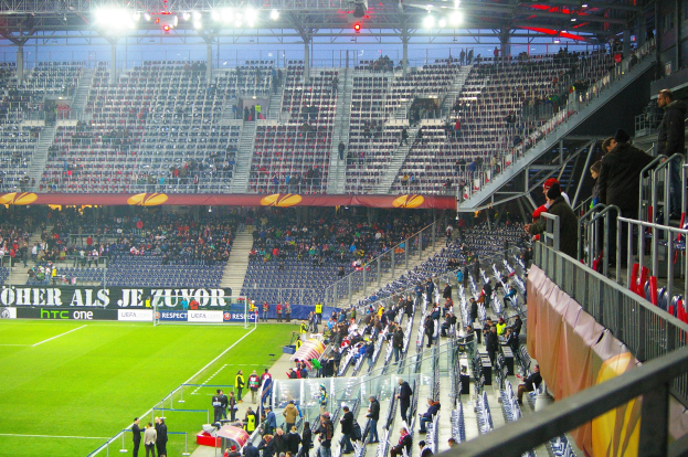 Ein großes Stadion mit Zuschauern bei einem Fußballspiel, mit sitzenden und stehenden Personen, einem Geländer, einer rechten Banner und einem leeren beleuchteten Bereich im Hintergrund.