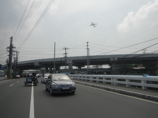 Ein Auto fährt auf einer Autobahn mit einer Brücke im Hintergrund, flankiert von Strommasten mit Oberleitungen, während ein Flugzeug am Himmel fliegt.