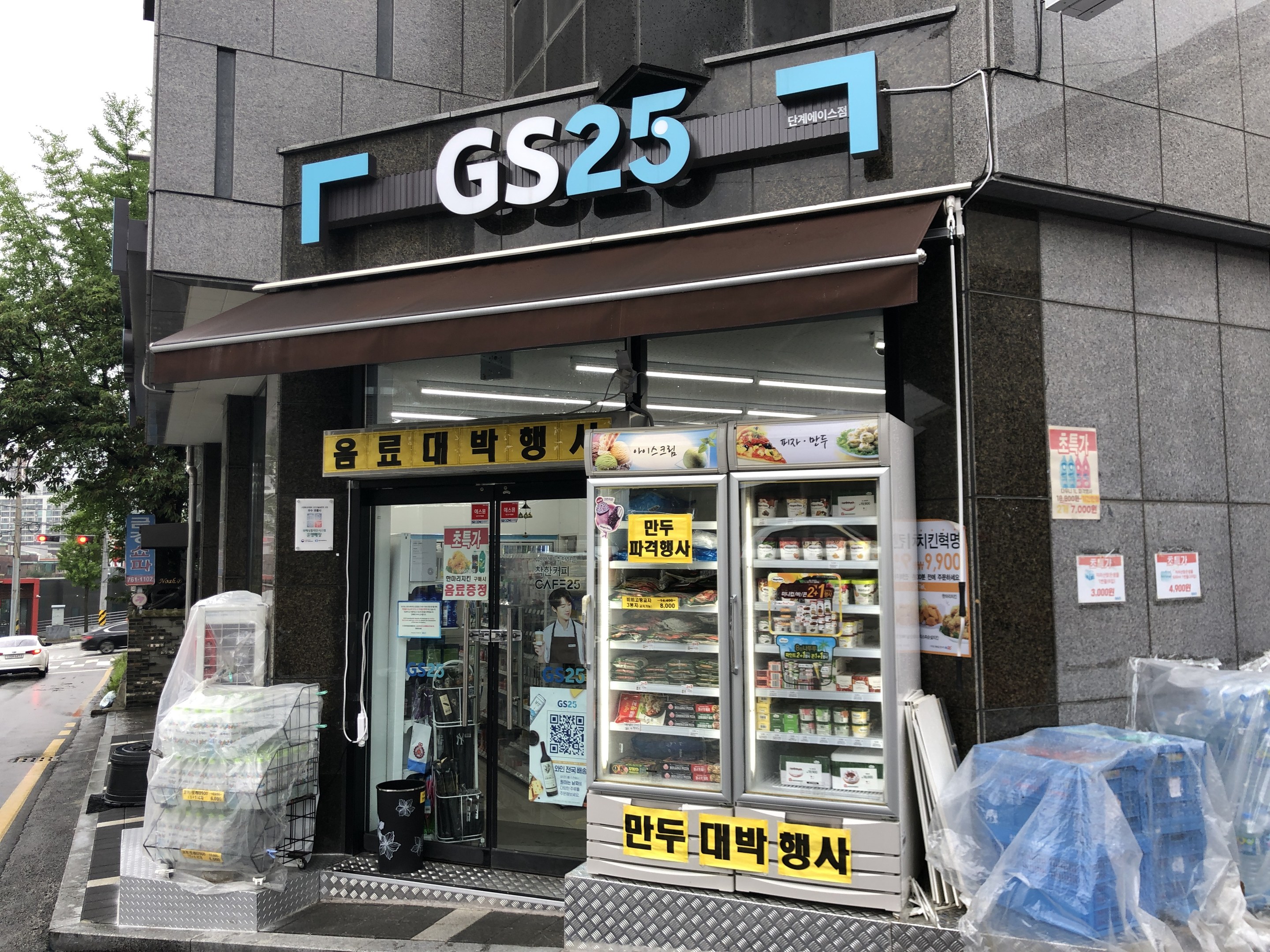 Ein Gebäude mit einer "GS25"-Reklame, umgeben von Fahrzeugen, Bäumen und einem klaren blauen Himmel, das einen koreanischen Lebensmittelmarkt mit Kühlschränken beherbergt, die mit Lebensmitteln gefüllt sind.