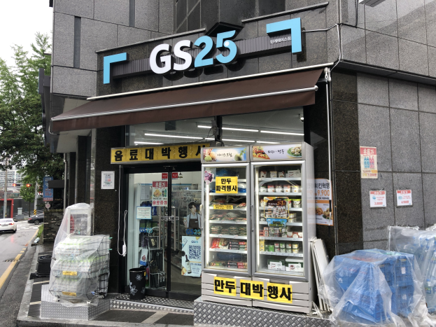 Ein Gebäude mit einer "GS25"-Reklame, umgeben von Fahrzeugen, Bäumen und einem klaren blauen Himmel, das einen koreanischen Lebensmittelmarkt mit Kühlschränken beherbergt, die mit Lebensmitteln gefüllt sind.