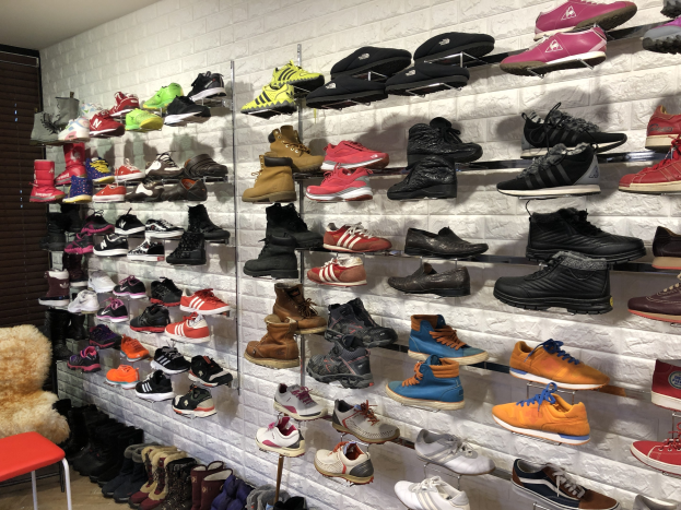 Ein Schuhgeschäftsinnenraum mit zahlreichen Schuhen auf Gestellen, einem Stuhl mit einem Teddybär links und einer Wand im Hintergrund.