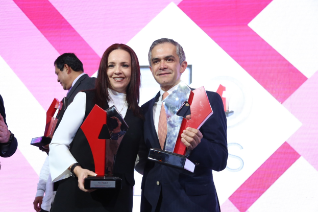 Zwei Personen auf einer Bühne, die strahlend Trophäen halten, mit einem Bildschirm im Hintergrund.