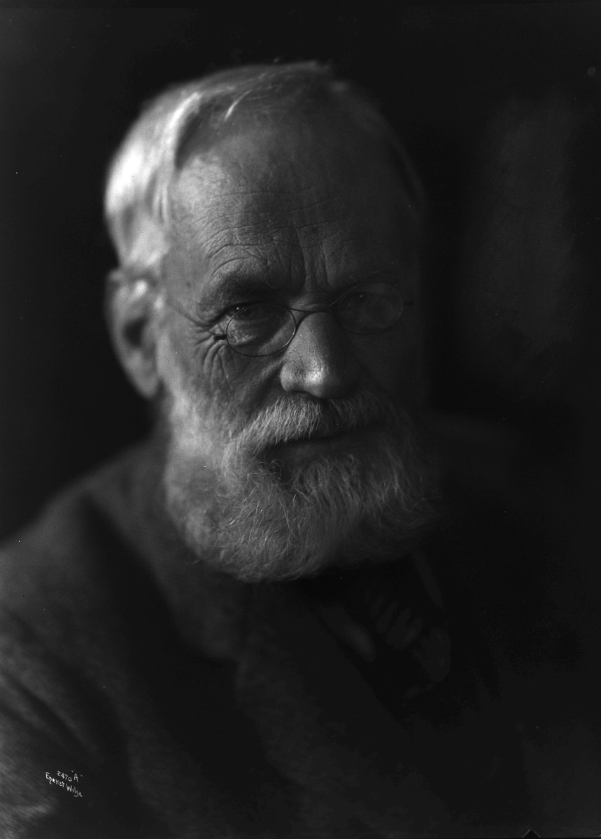 Alter Mann mit Vollbart und Brille, trägt dunklen Hintergrund, ernsthafter Ausdruck, konzentrierte Augen und ordentlich gekämmtes Haar.