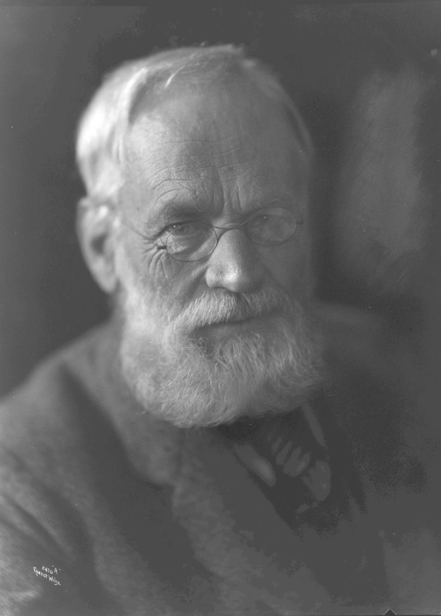 Alter Mann mit Vollbart und Brille, trägt dunklen Hintergrund, ernsthafter Ausdruck, konzentrierte Augen und ordentlich gekämmtes Haar.