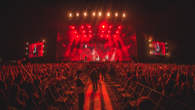 Eine große Menschenmenge steht vor einer hell erleuchteten Bühne mit Geländern, während Musiker spielen und andere zuschauen