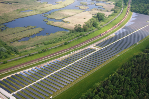 Luftaufnahme eines Solarparks mit Panelen, umgeben von B├Ąumen, Gras, Wasser und einer nahen Bahnschiene.