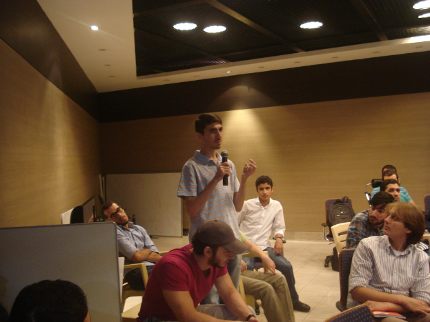 Eine Person steht in einem Büroraum und hält ein Mikrofon, während mehrere andere auf Stühlen sitzen.