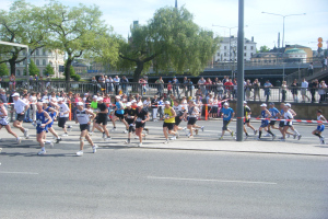 Gruppe von Läufern bei einem Marathon auf einer Straße mit Zielpfosten, Band, Absperrung, Pfosten, Schilder, Brücke, Gebäuden, Bäumen und bewölktem Himmel.