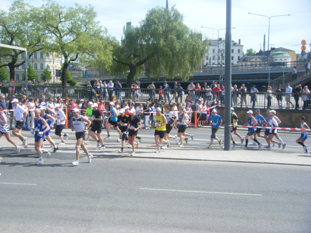Gruppe von Läufern bei einem Marathon auf einer Straße mit Zielpfosten, Band, Absperrung, Pfosten, Schilder, Brücke, Gebäuden, Bäumen und bewölktem Himmel.