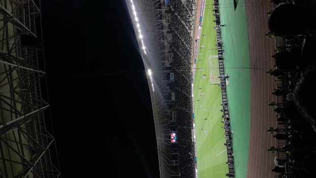 Ein Fußballspiel wird in einem Stadion bei Nacht gespielt, mit ein paar Menschen auf der rechten Seite, Metallstangen und Lichtern auf der linken Seite und einem dunklen Hintergrund, der eine dramatische Atmosphäre schafft.