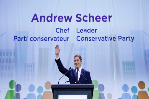 Andrew Scheer, der Vorsitzende der Konservativen Partei, steht hinter einem Podium mit zwei Mikrofonen und einem Bildschirm, mit einem Banner im Hintergrund mit Text und Gebäudeillustrationen.