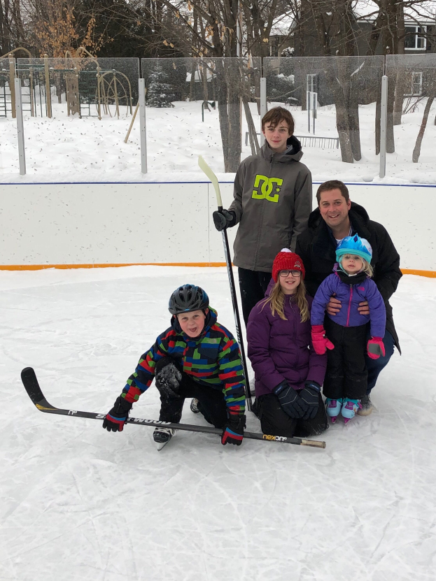 Eine Familie von vier Personen posiert auf einem Eisplatz: eine erwachsene Person in einer schwarzen Jacke mit einem Hockey-Schläger und zwei Kinder in lila Jacken und schwarzen Helmen, mit einer schneebedeckten Landschaft, Bäumen, Pfählen, einem Zaun, einem Gebäude und einem klaren Himmel im Hintergrund.