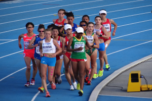 Eine Gruppe von Frauen, die auf einer Bahn laufen, mit einer Person auf der rechten Seite und einem gelben Objekt mit Drähten auf dem Boden, Tafeln mit Text im Hintergrund.