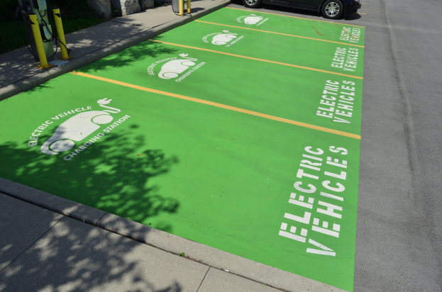 Eine Ladesäule für Elektrofahrzeuge in einer grünen Parkanlage mit einem daneben geparkten Auto, gelben Pfosten und einer Wand auf der linken Seite, einem weiteren Auto und einem Gebäude im Hintergrund.