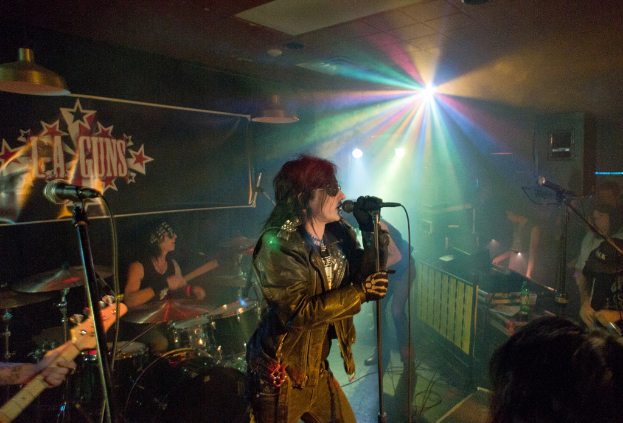 Eine Frau in einer schwarzen Jacke singt in ein Mikrofon auf der Bühne, mit einem Mann in Sonnenbrille und einem Headband, der ein Instrument spielt, im Hintergrund und einem Plakat mit der Aufschrift "LA Guns" im Hintergrund, unter Diskoleuchten und hängenden Lichtern.