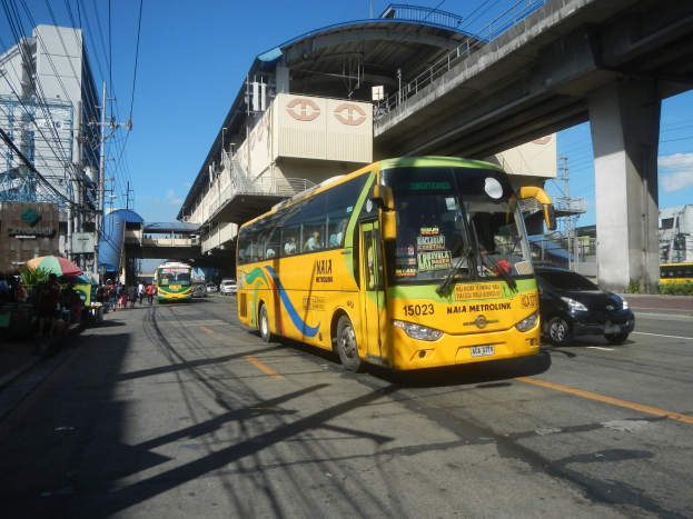 Ein gelber Bus fährt auf einer Stadtstraße mit hohen Gebäuden vorbei, Fußgänger auf dem Gehweg halten Regenschirme, eine Brücke mit Säulen und einer Überführung im Hintergrund, Strommasten mit Drähten und Texttafeln unter einem bewölkten Himmel.