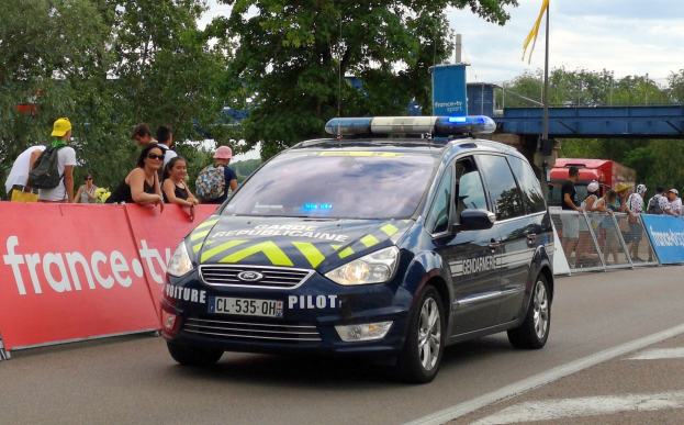 Polizeiauto fährt an einer Menge mit Schildern vorbei, Bäumen, einer Brücke, einer Flagge und einem bewölktem Himmel.