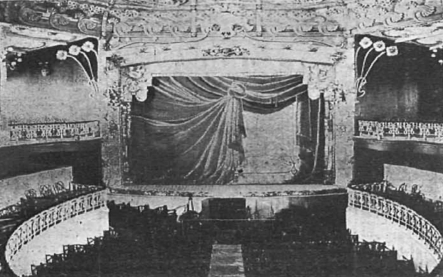 Schwarzes und weißes Foto des Theatersaals im Londoner Theatre Royal, das eine Bühne mit einem Vorhang, Geländern und kunstvollen Wanddesigns zeigt, sowie ein paar sitzende Menschen und Text am unteren Rand.