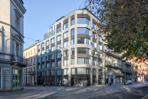 Großes modernes Bürogebäude mit zahlreichen Fenstern, umgeben von Straßeninfrastruktur, Fahrzeugen, Fußgängern, Bäumen und einem bewölkten Himmel, das als neuer Firmensitz dient.