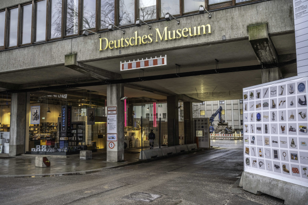 Außenansicht des Deutschen Museums in Berlin, das einen großen Gebäudekomplex mit Glasfenstern, Säulen und einem Namensschild zeigt, sowie ein Informationsschild mit anderen Gebäuden, Bäumen und Himmel im Hintergrund.
