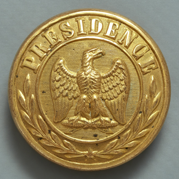 Eine goldene Münze mit einem Adler mit ausgebreiteten Flügeln und dem Wort "Präsident" in fetter Schrift, mittig auf einer flachen Oberfläche mit einem dekorativen Rahmen.