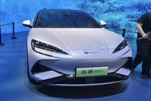 Ein weißes Auto auf einer Automesse mit einer Person, die rechts davon ein Objekt hält, ein Bildschirm im Hintergrund und Absperrpoller mit Seilen links.