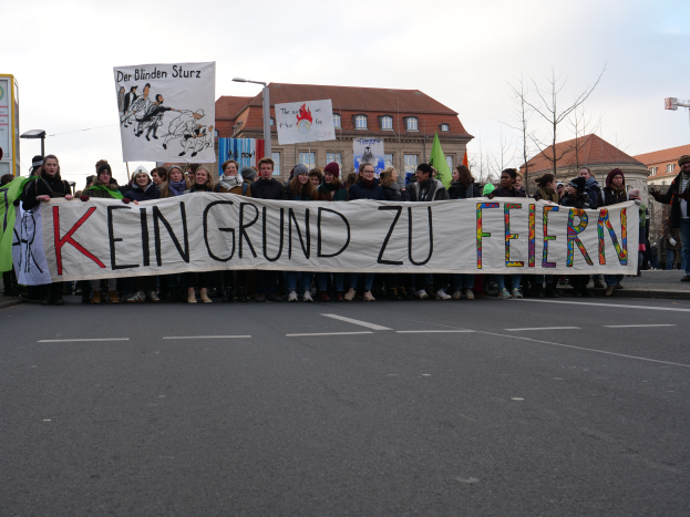 Eine Gruppe von Menschen auf der Straße mit einem Banner, auf dem "Kein Grund zu Feiern" steht, im Protest, mit Gebäuden, Laternenmasten, Bäumen und einem klaren blauen Himmel im Hintergrund.
