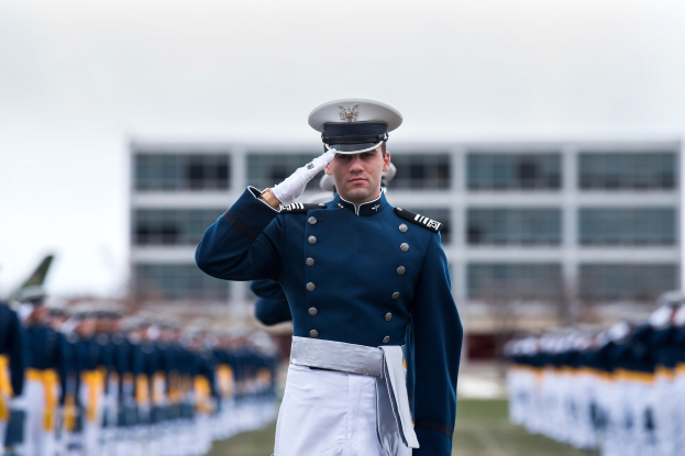 Ein Mann in militärischer Uniform salutiert auf einer Abschlussfeier umgeben von einer Menge, mit einem Gebäude und Himmel im Hintergrund.