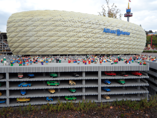 Eine große Lego-Struktur, die dem Allianz Stadion in München, Deutschland, ähnelt, mit einer Gruppe von Menschen davor, umgeben von Bäumen, Pfählen, einem Turm und Grünzeug unten, unter einem bewölkten Himmel.