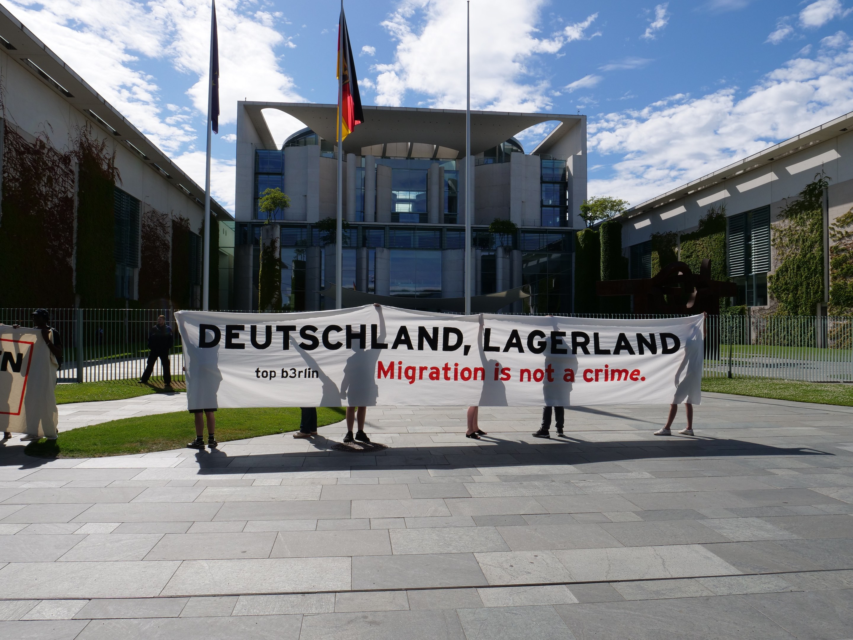 Gruppe von Menschen mit einem Banner mit der Aufschrift 'Deutschland, Lagerland Migration ist kein Verbrechen' vor einem Gebäude mit Fenstern, umgeben von Gras, einem Metallzaun, Pflanzen, Bäumen und Fahnenmasten mit Flaggen, unter einem bewölkten Himmel.