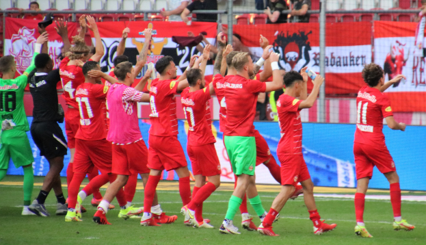 Gruppe von Fußballspielern, die ein Tor feiern, auf einem Feld, einige halten Flaschen oder klatschen, mit Bannern, einem Zaun, Stühlen und einer Decke mit Lampen im Hintergrund.