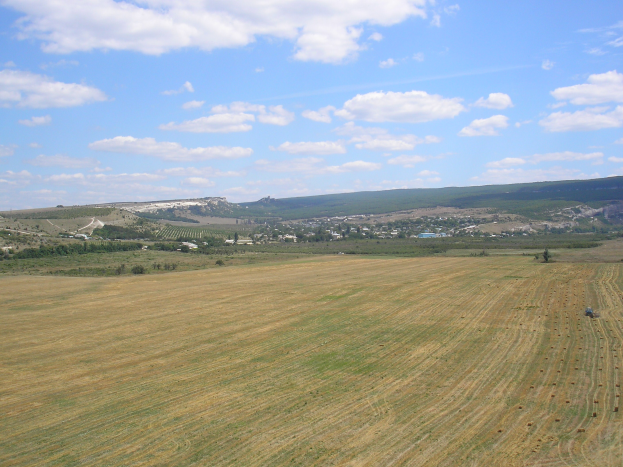 Luftaufnahme eines Traktors auf einem bewirtschafteten Feld umgeben von Bäumen, Gebäuden, Hügeln und einem bewölkten Himmel.