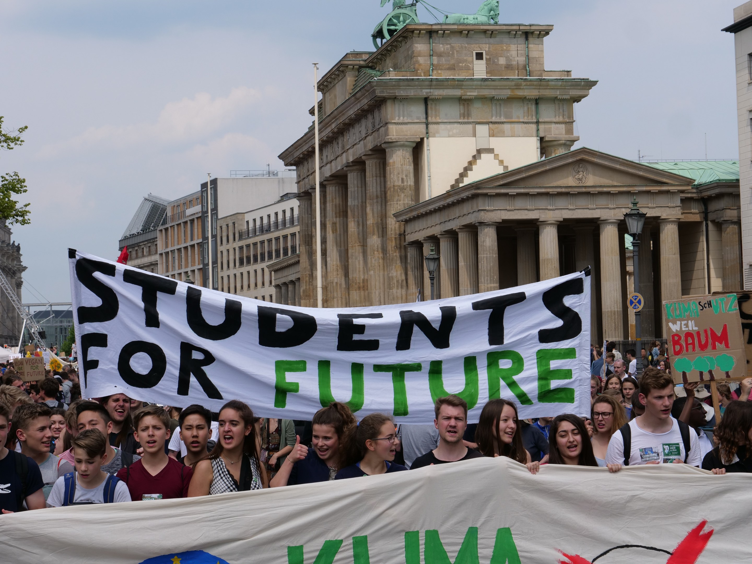 Schüler in Berlin marschieren mit einem leuchtend bunten "Students for Future"-Schild vor einem Hintergrund aus Gebäuden, Bäumen und Himmel.